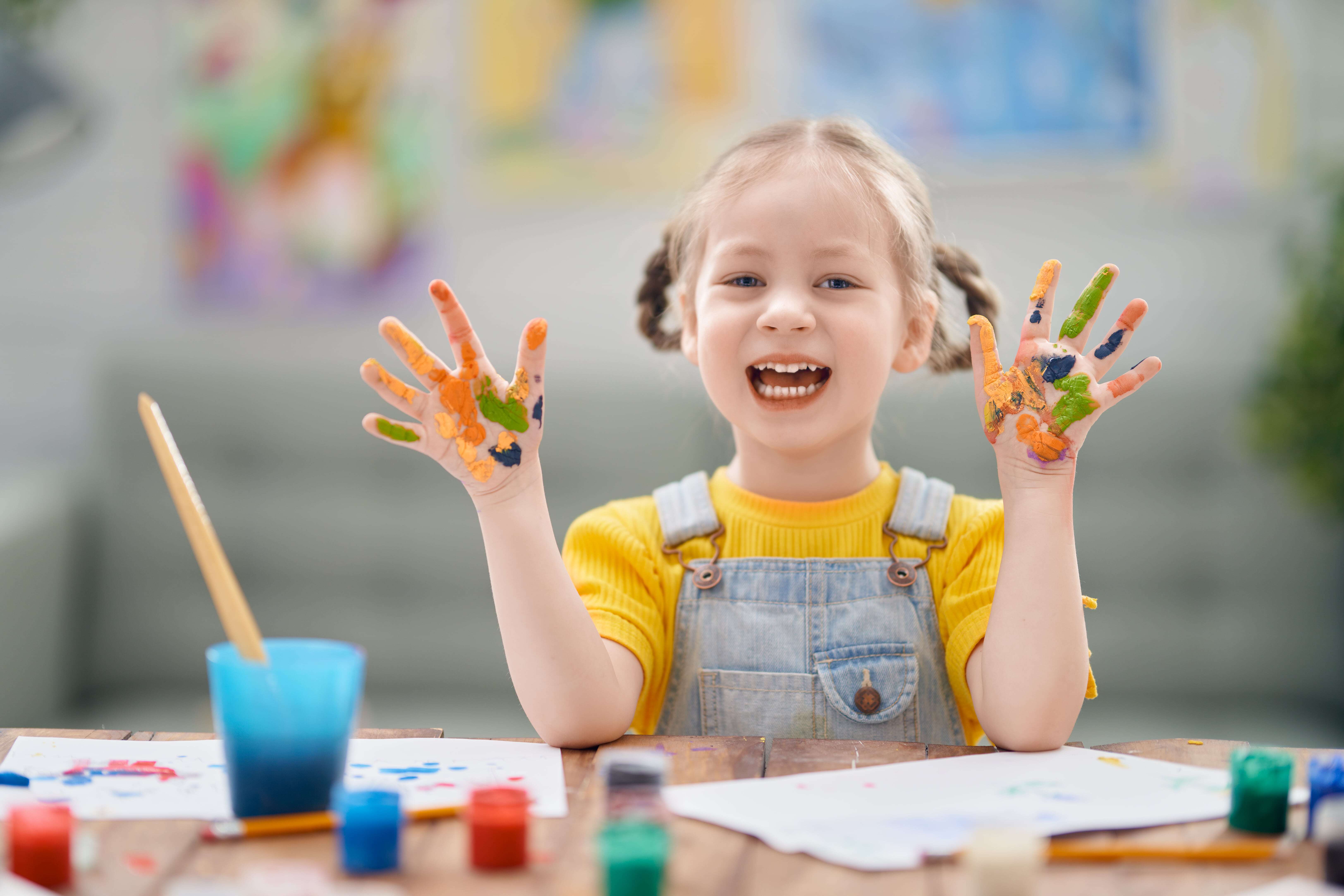 A young girl with paint on her hands, smiling and enjoying learning through play.
