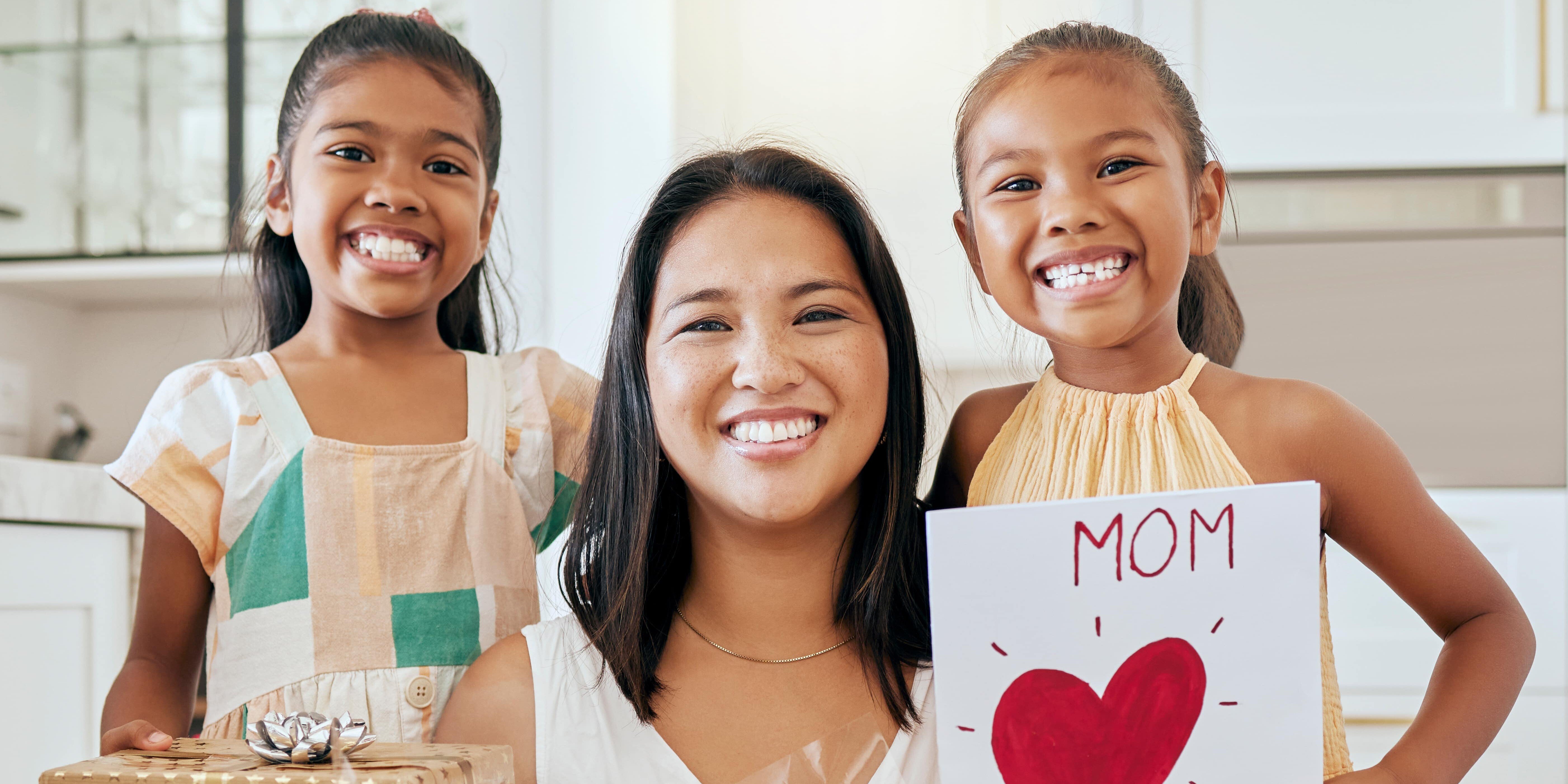 A mother holding flowers, kneeling in the middle of her two daughters who are holding a present and a card for Mother's Day.