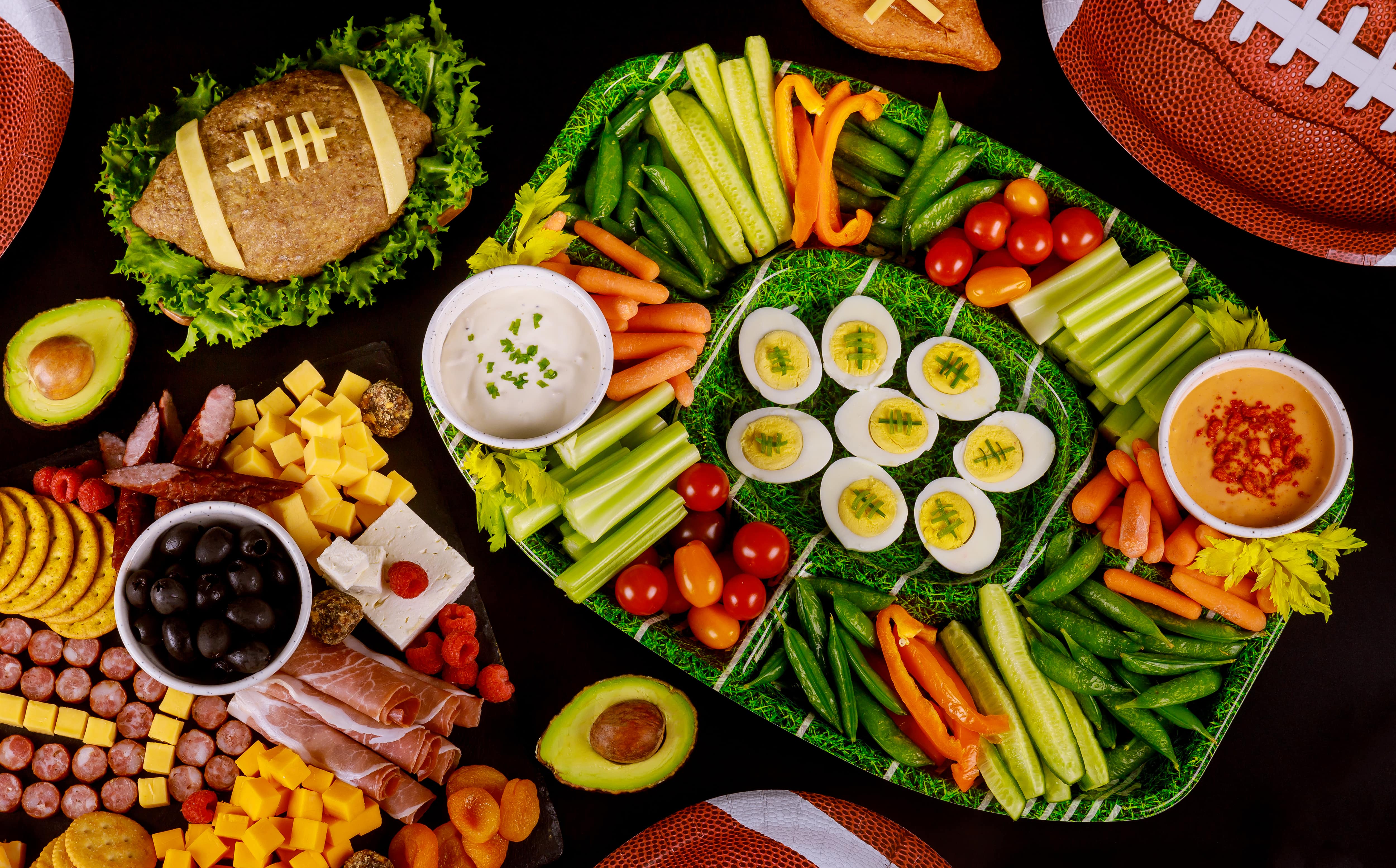 An overhead shot of healthy Super Bowl snacks for kids, including a veggie tray, an assortment of meats, and footballs for decoration. 