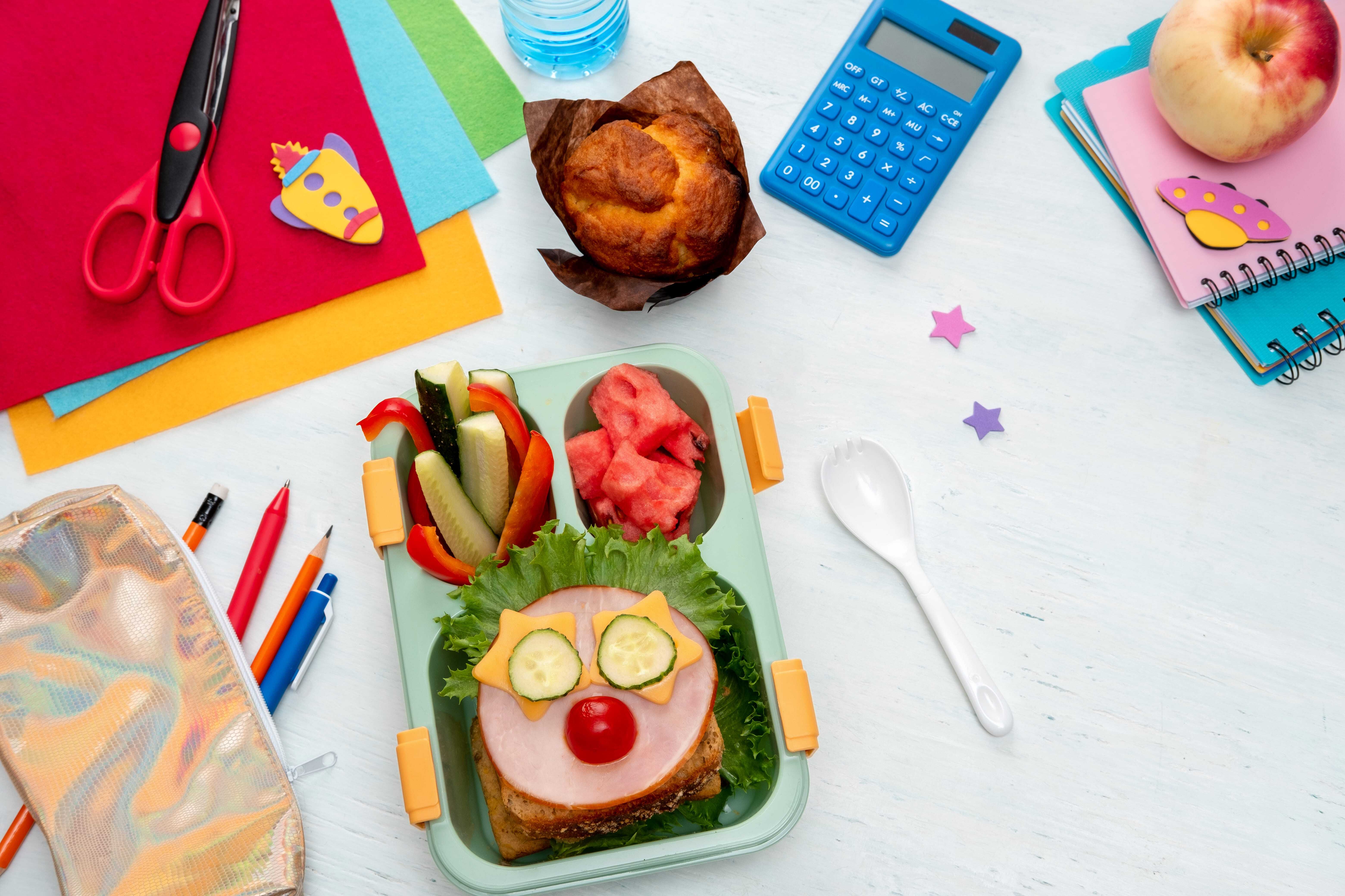 A child's lunch box with fruit, veggies, and a whimsical sandwich open on a table full of craft supplies. 