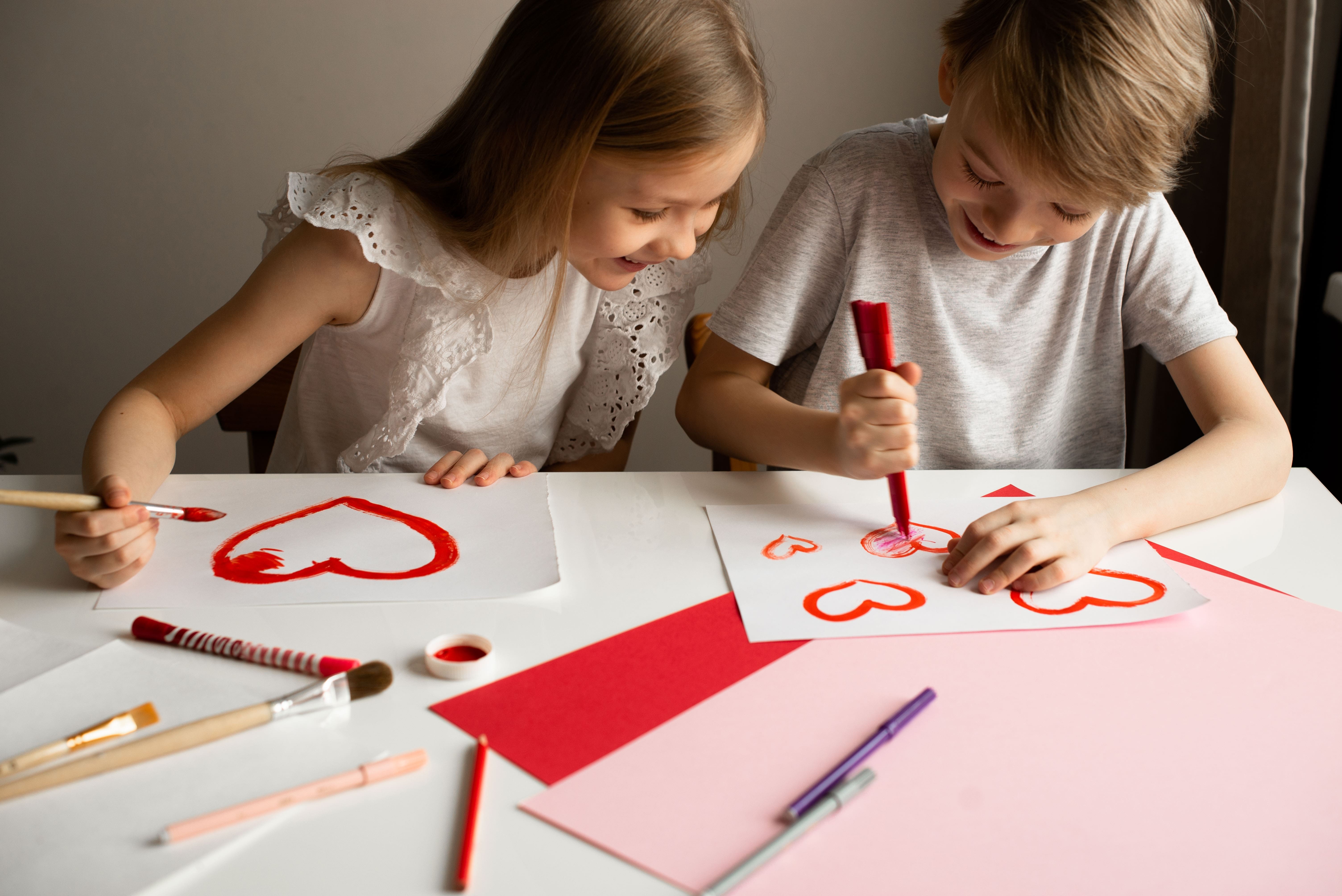 Two siblings sitting side-by-side drawing hearts on paper, making Valentine's Day crafts for kids