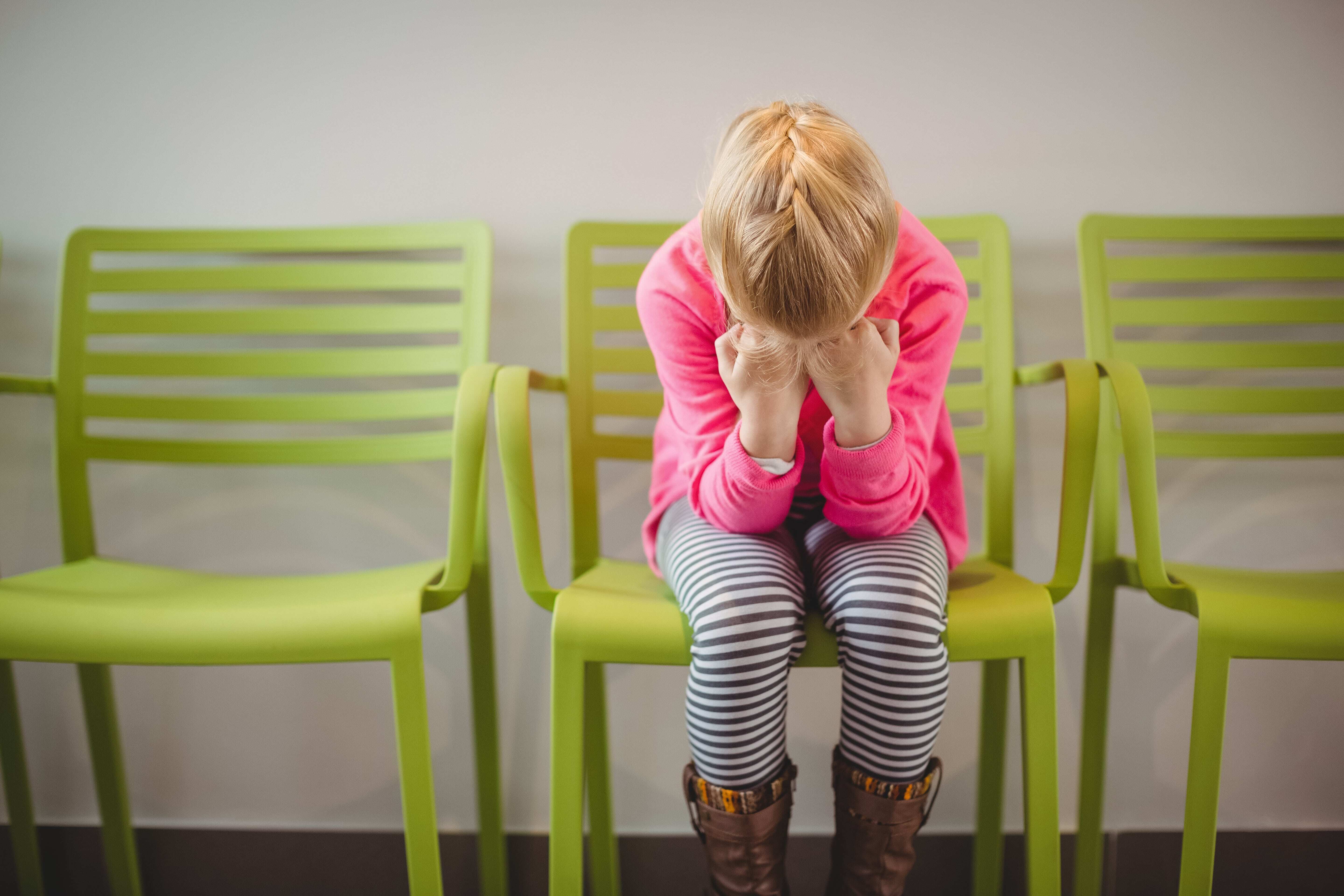 A young girl sitting in a green chair in the hallway of a school holding her head in her hands with feelings of back-to-school anxiety.